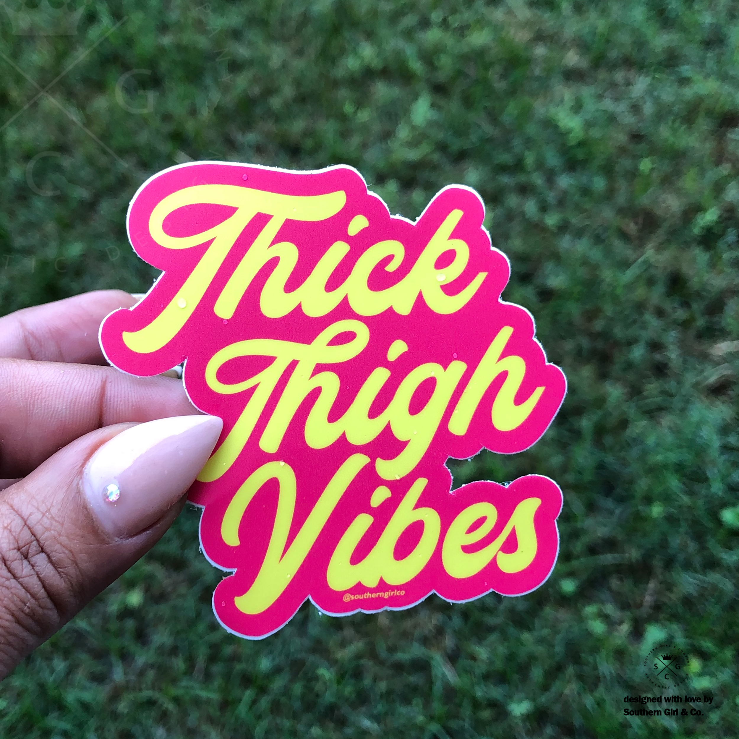 hand holding pink sticker with yellow writing that say Thick Thigh Vibes over grass